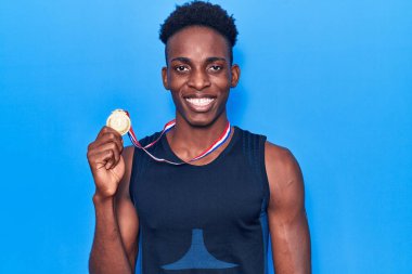 Afro-Amerikalı genç adam spor giysisi takıyor. Elinde madalya var. Olumlu ve mutlu görünüyor. 