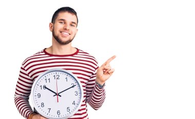 Büyük saati tutan yakışıklı genç adam gülümsüyor. Mutlu bir şekilde işaret ediyor. 