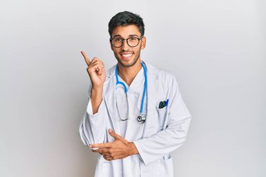 Young handsome man wearing doctor uniform and stethoscope with a big smile on face, pointing with hand and finger to the side looking at the camera. 