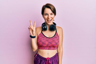 Kısa saçlı, spor giyimli, kulaklık takan, kameraya gülümseyen ve zafer işareti yapan esmer bir kadın. İki numara.. 