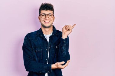 Gündelik kıyafetler ve gözlük takan, yüzünde kocaman bir gülümseme olan, eli ve parmağı kameraya bakan genç İspanyol bir adam.. 