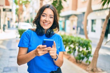 Genç İspanyol kadın şehirde akıllı telefon ve kulaklık kullanarak mutlu bir şekilde gülümsüyor..