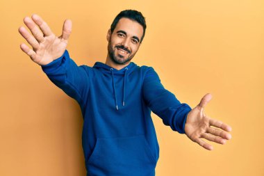 Gündelik giysiler giymiş genç İspanyol adam kameraya bakıp sarılmak için kollarını açmış gülümsüyor. Mutluluğu kucaklayan neşeli bir ifade.. 