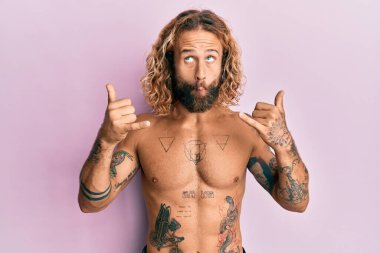 Handsome man with beard and long hair standing shirtless showing tattoos doing shaka sign making fish face with mouth and squinting eyes, crazy and comical. 