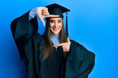 Beautiful brunette young woman wearing graduation cap and ceremony robe smiling making frame with hands and fingers with happy face. creativity and photography concept. 