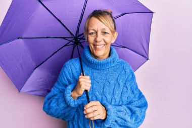 Orta yaşlı sarışın kadın mor şemsiye tutuyor. Pozitif ve mutlu görünüyor. Duruyor ve gülümsüyor. 