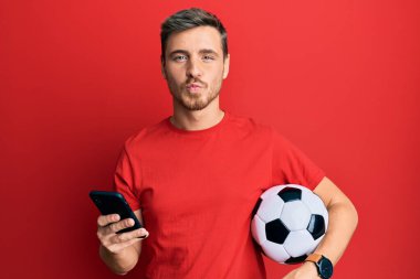 Yakışıklı, beyaz bir adam futbol topu tutuyor. Akıllı telefona bakıyor. Kameraya bakıyor. aşk ifadesi. 
