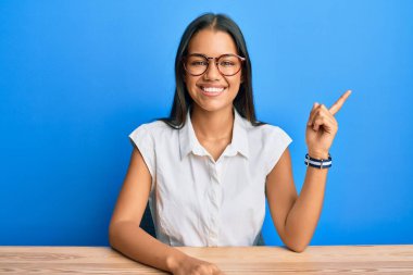 Güzel İspanyol kadın, gündelik giysiler içinde masada oturuyor, yüzünde kocaman bir gülümsemeyle, eli kameraya bakıyor.. 