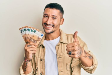 50 Euro 'luk banknotlar tutan yakışıklı adam mutlu ve olumlu gülümsüyor, başparmağıyla mükemmel ve onay işareti yapıyor. 