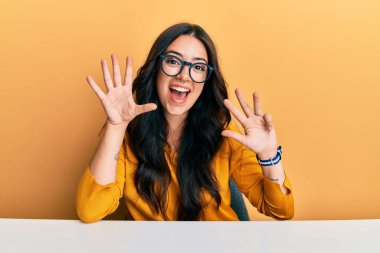 Güzel, esmer, gözlüklü ve gündelik giysileri olan genç bir kadın masanın üzerinde oturuyor ve dokuz numaralı parmaklarıyla işaret ediyor kendinden emin ve mutlu gülümsüyor.. 