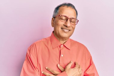 Orta yaşlı Hintli bir adam günlük kıyafetler ve gözlük takıyor elleri göğsünde, gözleri kapalı gülümsüyor ve yüzünde minnet ifadesi var. sağlık kavramı. 