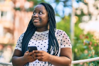 Genç Afrikalı Amerikalı kadın şehirde akıllı telefon kullanarak mutlu bir şekilde gülümsüyor..