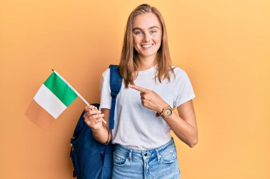 Güzel sarışın değişim öğrencisi İrlanda bayrağı taşıyor. Mutlu bir şekilde el ve parmak işareti yapıyor. 