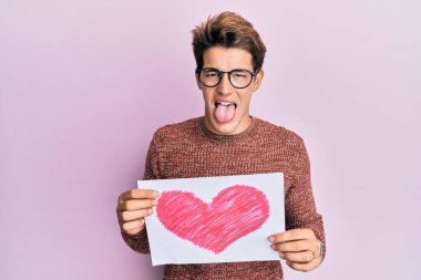 Handsome caucasian man holding heart draw sticking tongue out happy with funny expression. 