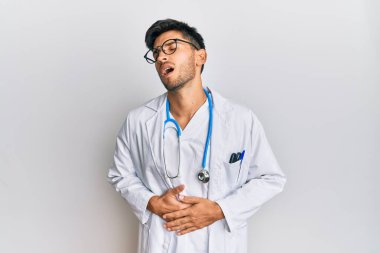 Young handsome man wearing doctor uniform and stethoscope with hand on stomach because nausea, painful disease feeling unwell. ache concept. 