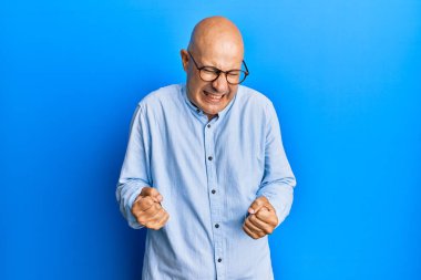 Orta yaşlı kel bir adam, günlük kıyafetler ve gözlük takıyor. Çok mutlu ve heyecanlı. Kolları havada, gülümseyen ve başarı için bağıran kazanan bir jest yapıyor. kutlama konsepti. 