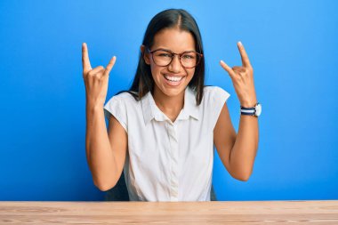 Güzel İspanyol kadın gündelik giysiler içinde masaya oturmuş deli gibi bağırıyor elleri havada rock sembolü yapıyor. Müzik yıldızı. ağır müzik konsepti. 