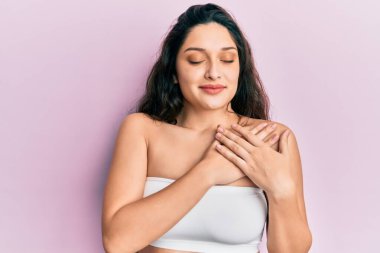 Beautiful middle eastern woman standing wearing a top showing skin smiling with hands on chest, eyes closed with grateful gesture on face. health concept. 