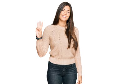 Kış süveteri giyen genç esmer kadın üç numaralı parmaklarını gösterip gülümserken kendinden emin ve mutlu.. 