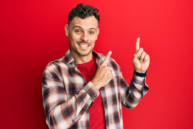 Gündelik kıyafetler giyen genç İspanyol adam gülümseyip kameraya bakıyor ve iki eliyle ve parmaklarıyla kenarı işaret ediyor.. 