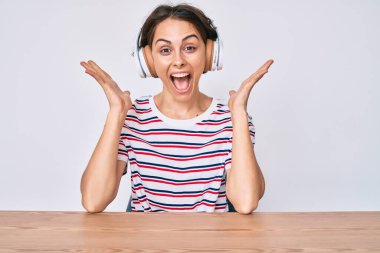 Masada oturmuş zaferi kutlayan kulaklık takan genç İspanyol kadın mutlu bir gülümseme ve ellerini kaldırarak kazanan yüz ifadesi takınıyor. 