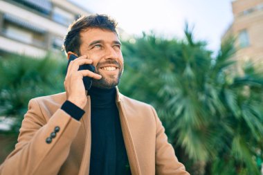 Zarif bir ceket giyen yakışıklı bir iş adamı akıllı telefon kullanarak dışarıda mutlu bir şekilde gülümsüyor.