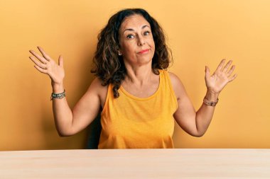 Orta yaşlı, sıradan kıyafetler giyen güzel bir kadın masanın üzerinde elleri ve kolları havada, kafası karışmış bir şekilde oturuyor. şüphe kavramı. 