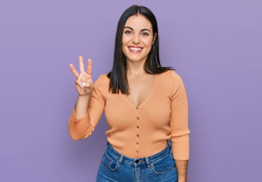 Genç İspanyol kadın gündelik kıyafetler giyer ve üç numaralı parmaklarıyla gülümseyerek mutlu ve kendinden emin bir şekilde işaret eder.. 