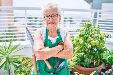 Gri saçlı, eldivenli ve bahçıvan önlüklü yaşlı kadın evdeki bitkileri bahçeliyor.
