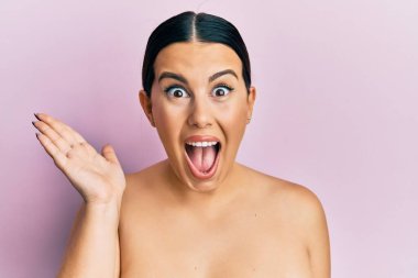 Güzel esmer kadın üstsüz duruyor başarıyı kutlayan tenini gösteriyor mutlu bir gülümsemeyle ve elini kaldırarak kazanan yüz ifadesiyle. 