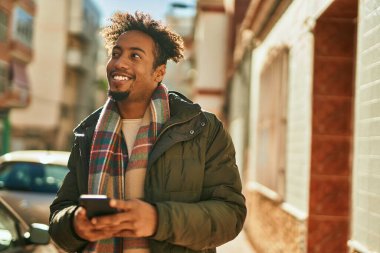 Genç Afrikalı Amerikalı adam şehirde akıllı telefon kullanarak mutlu bir şekilde gülümsüyor.