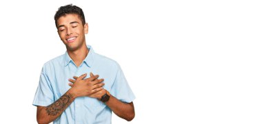 Genç, yakışıklı, Afro-Amerikalı sıradan kıyafetler giyen, elleri göğsünde, gözleri kapalı gülümseyen ve yüzünde minnet ifadesi olan bir adam. sağlık kavramı. 