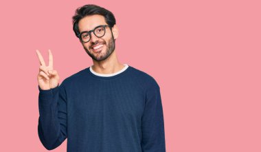 Gündelik kıyafetler giyen ve gözlüklü genç İspanyol adam iki numarayı gösterip işaret ederken kendinden emin ve mutlu bir şekilde gülümsüyor.. 