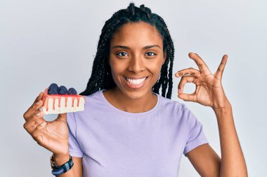 Elinde bir dilim peynirli kek tutan Afro-Amerikalı kadın parmaklarıyla tamam işareti yapıyor, gülümsüyor. 