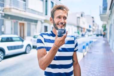 Sakallı yakışıklı beyaz adam dışarıda mutlu bir şekilde gülümsüyor telefonda sesli mesaj gönderiyor.