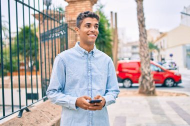 Genç Latin adam mutlu bir şekilde gülümsüyor ve şehirde akıllı telefon kullanıyor..