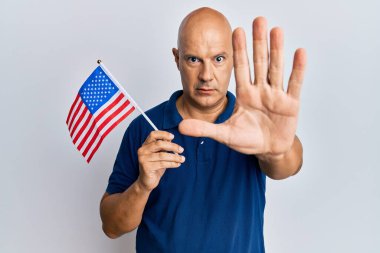 Orta yaşlı, kel bir adam elinde bayrakları açık bir şekilde tutuyor dur işareti yapıyor ciddi ve kendinden emin bir ifadeyle, savunma hareketi yapıyor. 