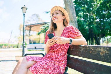 Genç sarışın kadın tatilde. Parktaki bankta oturan akıllı telefonu kullanıyor.