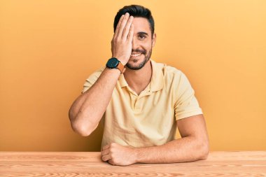 Latin asıllı genç adam gündelik giysiler içinde masada oturuyor bir gözünü eliyle kapatıyor, yüzünde kendine güvenen bir gülümseme ve sürpriz bir duygu var.. 