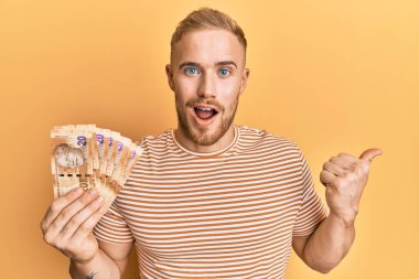 Güney Afrika 'da elinde 20 rand banknot tutan beyaz bir adam başparmağıyla yan tarafa bakıyor ve ağzı açık gülümsüyor. 