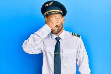 Sakallı, uçak pilotu üniforması giyen, gülümseyen ve elleri yüzünde, şaşırtmak için gözlerini kapatan yakışıklı bir adam. kör kavram. 