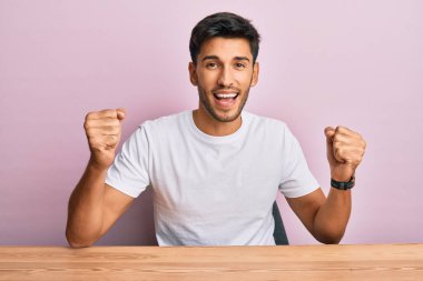Genç yakışıklı adam, gündelik beyaz tişört giyip masada oturmuş gururla bağırıyor zaferi ve başarıyı kutluyor. Kollarını kaldırıp heyecanla bekliyor. 