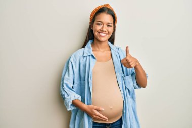 Güzel İspanyol bir kadın bebek bekliyor, hamile karnına dokunuyor mutlu bir el hareketi yapıyor. Kameraya bakıp başarıyı gösteren ifade. 