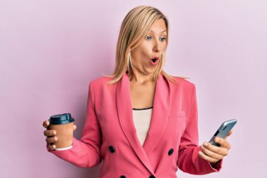 Güzel, orta yaşlı, sarışın bir kadın akıllı telefon kullanıyor ve bir fincan kahve içiyor. Korkmuş ve şok olmuş bir şekilde şaşkınlık, şaşkınlık ifadesi, korku ve heyecanlı bir yüz ifadesi var.. 