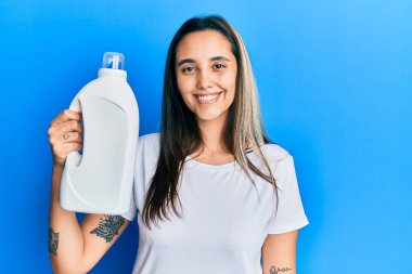 Deterjan şişesi tutan genç İspanyol kadın pozitif ve mutlu görünüyor. Dişlerini gösteren kendine güvenen bir gülümsemeyle ayakta duruyor ve gülümsüyor. 