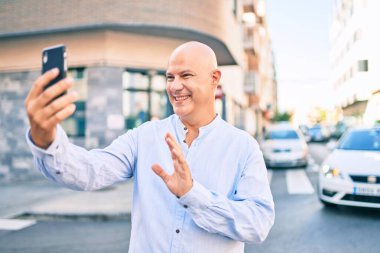 Orta yaşlı kel bir adam şehirde akıllı telefon kullanarak video araması yaparken mutlu gülümsüyor..