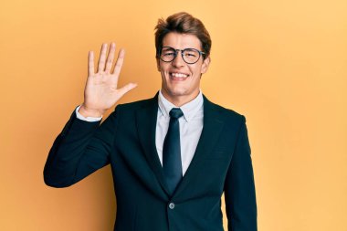 Yakışıklı, beyaz bir adam, iş elbisesi ve kravat takıyor. Beş numaralı parmaklarıyla işaret ediyor ve gülümsüyor. Kendinden emin ve mutlu.. 