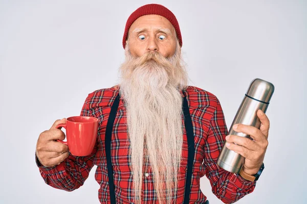 Old senior man with grey hair and long beard holding cup of coffee and thermo making fish face with mouth and squinting eyes, crazy and comical.