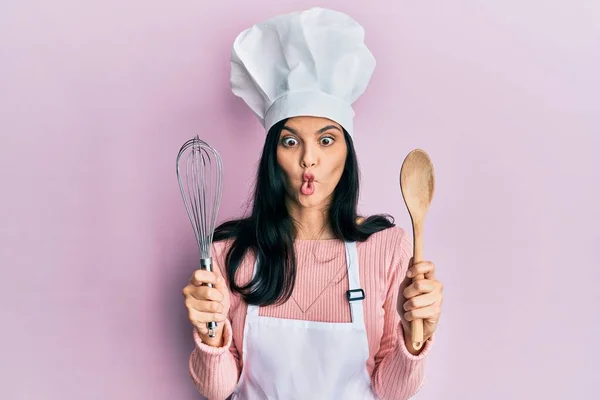 Young hispanic woman wearing baker uniform holding spoon and whisk making fish face with mouth and squinting eyes, crazy and comical.