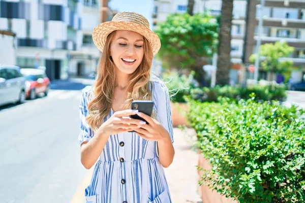 年轻美丽的金发女子戴着夏帽在度假时快乐地笑着 在城市街道上用智能手机笑着站在那里 — 图库照片